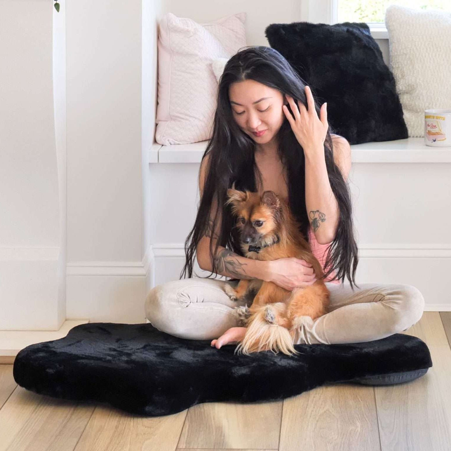 A person sitting on the floor with a small brown dog on a black luxury faux fur dog bed, which is leakproof and washable.